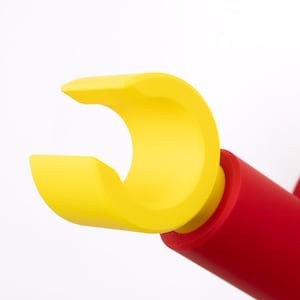 Op de afbeelding: Een heldergele, gebogen, open vorm is bevestigd aan een rode cilindrische basis. Het ontwerp doet denken aan een hand of een grijpgereedschap. Het object is op een wit oppervlak gemonteerd.