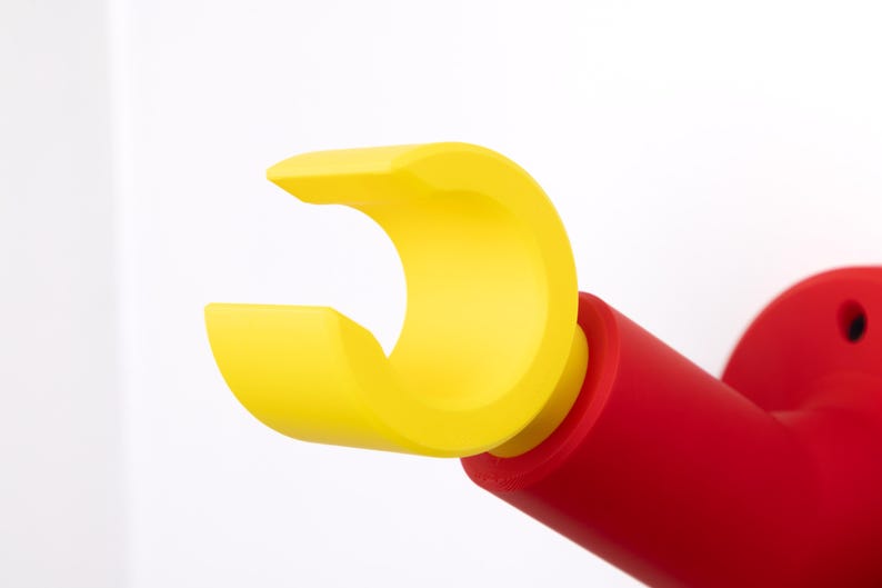 Op de afbeelding: Een heldergele, gebogen, open vorm is bevestigd aan een rode cilindrische basis. Het ontwerp doet denken aan een hand of een grijpgereedschap. Het object is op een wit oppervlak gemonteerd.
