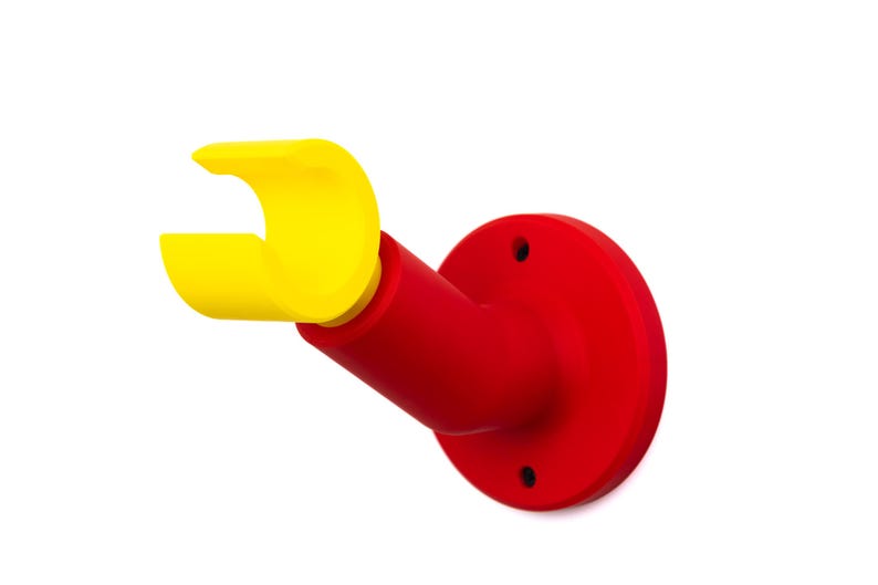 Op de afbeelding: Een aan de muur gemonteerde beugel in heldere primaire kleuren. De basis is helderrood, gaat over in een rode schuine arm en eindigt in een gele, gebogen houder. Het ontwerp is eenvoudig en modern, met een strakke, minimalistische esthetiek.