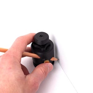May include: A black circle cutter tool with a pencil inserted, held by a hand. The pencil is used to draw a line on a white surface. The tool has a knob for adjusting the cutting radius.