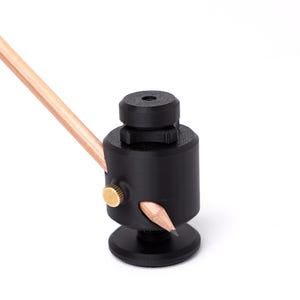 May include: A black pencil sharpener with a gold-colored screw and a pencil inserted. The pencil is light brown. The sharpener has a cylindrical body and a round base. The background is white.