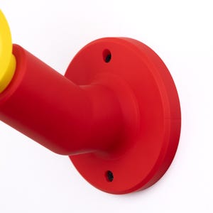 Op de afbeelding: Een rood en geel aan de muur bevestigd object met een cirkelvormige basis en een cilindrische arm. Het object is gemaakt van een glad, stevig materiaal en staat tegen een witte achtergrond. Het gele gedeelte is een cirkelvormige opening.