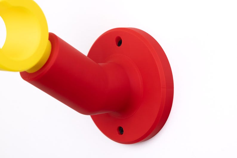 Op de afbeelding: Een rood en geel aan de muur bevestigd object met een cirkelvormige basis en een cilindrische arm. Het object is gemaakt van een glad, stevig materiaal en staat tegen een witte achtergrond. Het gele gedeelte is een cirkelvormige opening.