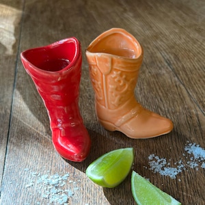 May include: Two ceramic shot glasses shaped like cowboy boots, one red and one orange, sit on a wooden surface. Two lime wedges and a scattering of salt are also present, suggesting a tequila shot setup.