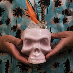 May include: A pink ceramic skull mug with a tropical flower garnish and ice cubes. The mug is being held by a person wearing a blue and white patterned shirt.