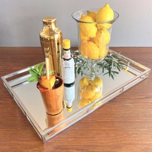 Könnte beinhalten: Ein verspiegeltes Serviertablett mit silbernem Rahmen enthält einen Cocktail, einen goldenen Cocktailshaker und eine Glasvase voller Zitronen. Eine Flasche Bitter und ein Minzzweig befinden sich ebenfalls auf dem Tablett. Der Cocktail befindet sich in einem Holzbecher mit Ananas-Garnitur.