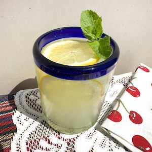 Puede incluir: Un vaso transparente con un borde grueso de color azul cobalto que contiene un líquido amarillo pálido, dos rodajas de limón flotando en su interior y una ramita de menta verde fresca decorando el borde superior.