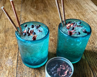 May include: Two turquoise blue glasses filled with ice, dried flower petals, and dark blue berries. The glasses have copper straws and are on a wooden table. A small silver tin with dried flower petals and berries is in the foreground.