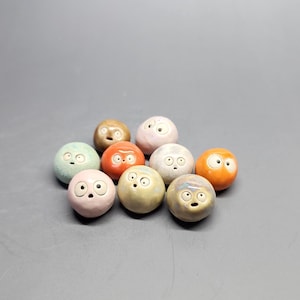 May include: A collection of nine small, spherical ceramic objects, each featuring a unique glazed finish in colors including pink, orange, blue, tan, and grey. Every sphere has two small, wide-set eyes and a tiny, open mouth, creating a facial expression.