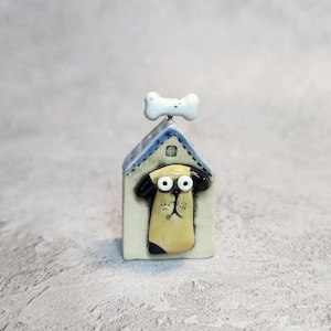 May include: A small ceramic figurine of a tan dog with black ears peeking out of a house-shaped structure. The house has a blue roof with a white bone-shaped topper. The dog has large white eyes and a black nose, set against a cream background.