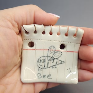 May include: A ceramic notepad-shaped trinket with a hand-drawn bee illustration and the word "Bee". The off-white piece has red lines and a perforated top edge, with a smiley face drawn on the bottom right.