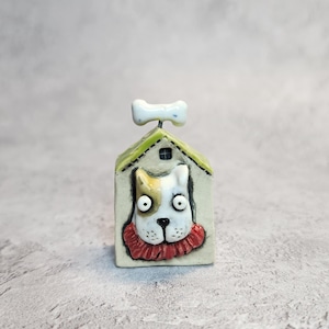 May include: A small ceramic figurine of a dog house with a white, brown, and black dog face peeking out. The dog wears a red ruffled collar. The house has a light green roof and a white bone-shaped ornament attached to the top peak.