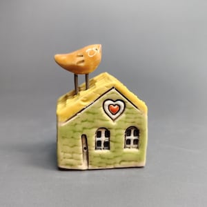 May include: A ceramic house figurine with a yellow roof and green walls. A small orange bird sits atop the roof. The house has two windows, a door, and a heart-shaped design. The figurine is a charming decorative item.