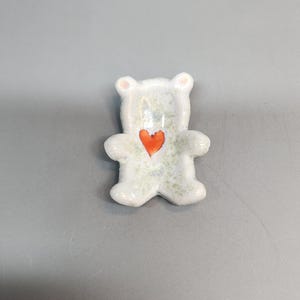 Puede incluir: Pequeña figura de oso de cerámica con un corazón rojo en el pecho. El oso es blanco con sutiles motas verdes y detalles rosas en las orejas. La figura mide unos 5 cm de altura.