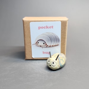 May include: A small, pale yellow, smooth, rounded figurine with two black antennae and large eyes, positioned in front of a square brown cardboard box featuring a white label with the text pocket bug and an illustration of a grey pill bug.