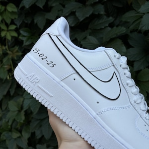 May include: White leather low-top sneakers with a black outlined Nike swoosh and the date "25-02-25" written on the side.