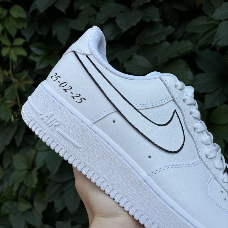 May include: White leather low-top sneakers with a black outlined Nike swoosh and the date "25-02-25" written on the side.