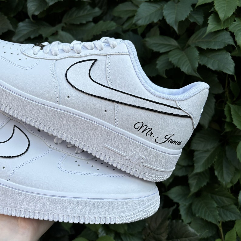 May include: White leather sneakers with a black Nike swoosh and the text "Mr. James" written on the side.