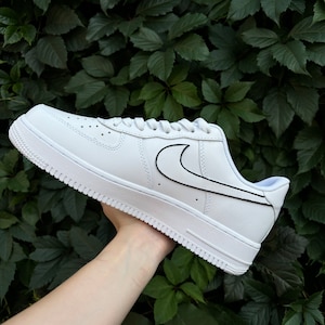 May include: White leather low-top sneakers with a black outline around the Nike swoosh logo.