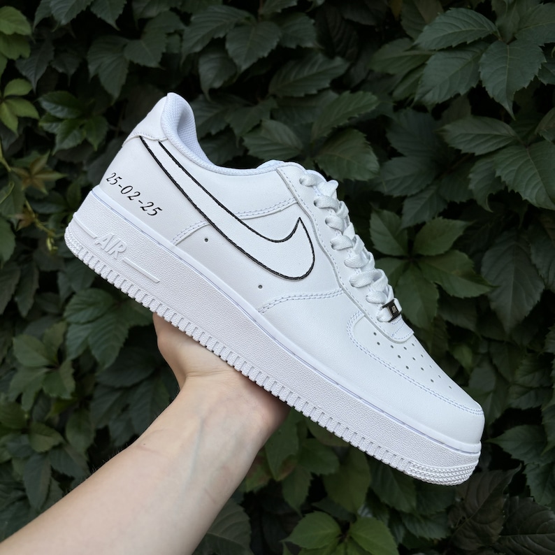 May include: White leather low-top sneakers with a black outlined Nike swoosh and the date "25-02-25" written on the side.