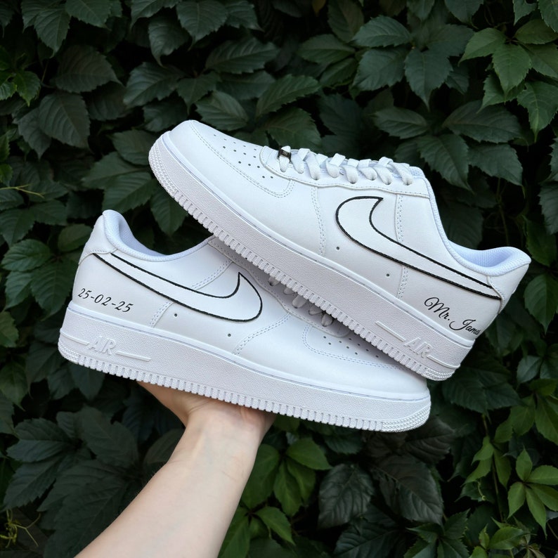 May include: A pair of white leather sneakers with black outlines around the Nike swoosh and text. One shoe has the text "Mr. James" and the other has the text "23-02-23".