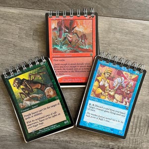 May include: Three Magic: The Gathering cards, each with a different creature depicted. The first card is green and features a deadly insect. The second card is red and features a lightning hound. The third card is blue and features a balloon peddler.