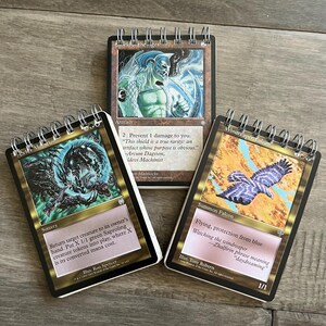 May include: Three Magic: The Gathering cards, each with a gold border and a different illustration. The first card is "Baleld the Geol", an artifact with a blue and green illustration. The second card is "Erft Mutation", a sorcery with a black and green illustration. The third card is "Windreaper Falcon", a summon falcon with a blue and brown illustration.