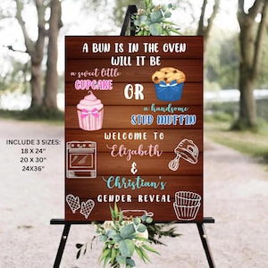 May include: A wooden sign with a brown background and white text announcing a gender reveal party for Elizabeth and Christian. The sign says "A bun is in the oven, will it be a sweet little cupcake or a handsome stud muffin?"  The sign includes a pink cupcake and a blue muffin with a bow on top. The sign also includes a picture of a mixer and an oven.