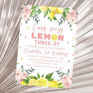 May include: A lemon-themed birthday invitation featuring pink flowers, lemons, and green leaves. The text reads "Easy peezy LEMON THREE-ZY. Please join us to celebrate Elizabeth's 3rd Birthday! Saturday | November 24th | 3PM. Event Center Anywhere Street. Any City Avenue 12345. RSVP to Michelle at 123-456-7890."