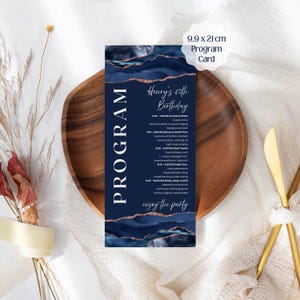 以下が含まれることがあります： ネイビーブルーのプログラムカード、9.9 x 21 cm、金色のアクセントと「Program」の文字。カードはヘンリーの60歳の誕生日用で、詳細なスケジュールが記載されています。カードは、金色のナイフが付いた木製のプレートの上にあります。