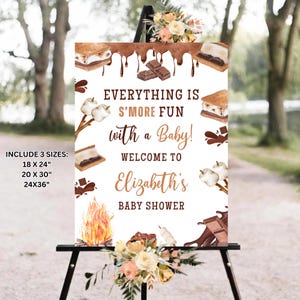 May include: A white sign with brown text that says "Everything is S'more Fun with a Baby! Welcome to Elizabeth's Baby Shower". The sign is decorated with images of s'mores, chocolate, and a campfire.