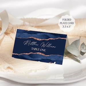 以下が含まれることがあります： ネイビーブルーの水彩デザインとローズゴールドのグリッターアクセントが施された、折り畳まれた席札、8.9 x 5.1 cm。カードには「Matthew Williams」と「TABLE ONE」という名前がエレガントな筆記体で書かれています。カードは白いお皿の上に表示されています。
