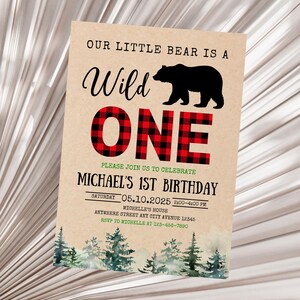 May include: A rustic brown invitation with a red and black plaid design. The text reads "Our little bear is a Wild One" with a black silhouette of a bear. The invitation is for Michael's 1st birthday party on Saturday, 05.10.2025 from 2:00-4:00 PM at Michelle's house. The address is Anywhere Street Any City Avenue 12345. RSVP to Michelle at 123-456-7890.