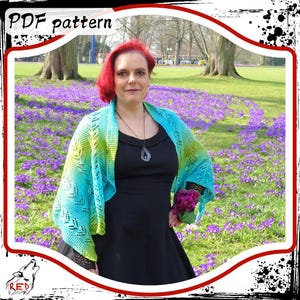 May include: A woman wearing a black dress and a teal, green, and blue crocheted shawl. She is standing in a field of purple flowers. The shawl has a delicate lace pattern.