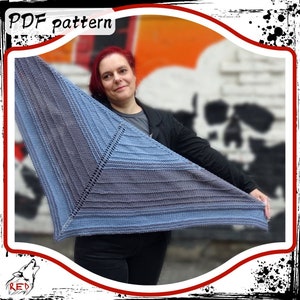 Puede incluir: Un chal de punto azul y gris con un patrón geométrico. El chal está siendo sostenido por una persona frente a una pared con grafitis.