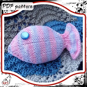 Può includere: Un pesce di lana rosa e viola con un occhio di bottone blu. Il pesce è sdraiato su un tessuto di lana blu e grigio.