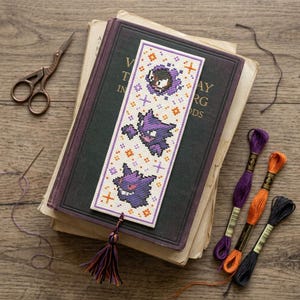 Könnte beinhalten: Ein rechteckiges Kreuzstich-Lesezeichen mit drei vertikal angeordneten, violetten, geisterartigen Kreaturen, die mit kleinen orangefarbenen und violetten Sternmotiven verziert sind. Das Lesezeichen endet in einer mehrfarbigen Quaste in Violett, Orange und Schwarz.