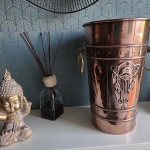 Peut inclure: Un seau en cuivre poli doté de deux poignées en anneau en laiton sur les côtés et d'un motif floral en relief sur le devant. Le récipient cylindrique présente un bord et une base renforcés avec des rivets visibles, offrant une finition métallique.