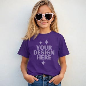 May include: A young person wearing a purple t-shirt with the text "YOUR DESIGN HERE" in white. The person is also wearing heart-shaped sunglasses and blue jeans. The background is a plain white wall.
