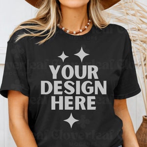 May include: A black t-shirt with the text "YOUR DESIGN HERE" in white. The shirt is being worn by a person with long blonde hair and a brown hat.