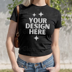 以下が含まれることがあります： 黒いクロップドTシャツは、短い袖と前面に白で「YOUR DESIGN HERE」という文字が印刷されています。シャツは、茶色のハンドバッグを持った人が着用しています。