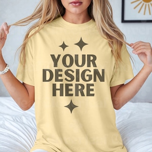 May include: A pale yellow t-shirt with short sleeves and a crew neck. The shirt has a design on the front that says "YOUR DESIGN HERE" with four small stars around the text.