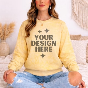Może przedstawiać: Jasnożółta bluza z napisem "YOUR DESIGN HERE" w ciemnoszarym kolorze. Bluza ma okrągły dekolt i długie rękawy. Modelka ma na sobie podarte niebieskie dżinsy. Tło obejmuje poduszki i elementy dekoracyjne.