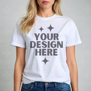 Puede incluir: Una camiseta blanca con el texto "YOUR DESIGN HERE" en gris, junto con tres gráficos de estrellas grises. La camiseta es de manga corta y está hecha de un material suave. La modelo lleva vaqueros azules.