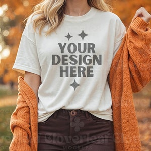 May include: A white t-shirt with the text "YOUR DESIGN HERE" in gray lettering and two small stars on either side of the text. The shirt is being worn by a person in a fall setting with orange leaves in the background.
