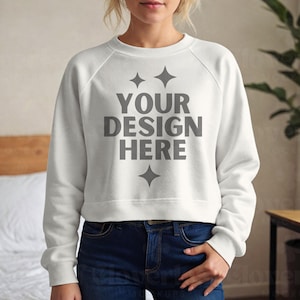 May include: A white sweatshirt with the text "YOUR DESIGN HERE" in gray, along with star graphics. The sweatshirt has long sleeves and a crew neck. The person is wearing blue jeans.