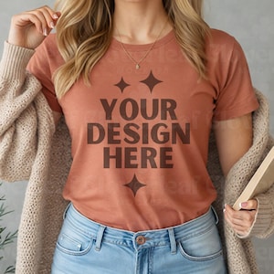 May include: A woman wearing a rust-colored t-shirt with the text "YOUR DESIGN HERE" in brown lettering. The shirt has a simple design with two small stars above the text and one below. She is wearing blue jeans and a brown cardigan.