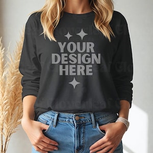 May include: A black long-sleeve shirt with the text "YOUR DESIGN HERE" in gray lettering. The shirt is worn by a person with long blonde hair and blue jeans.