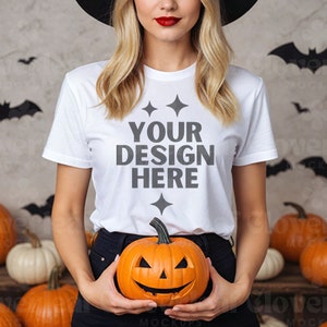 May include: A white t-shirt with the text "YOUR DESIGN HERE" in grey lettering. The shirt is being worn by a person holding a carved orange pumpkin. The background is a textured wall with black bats.