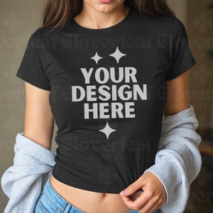 Op de afbeelding: Een zwart T-shirt met een wit ontwerp met de tekst "YOUR DESIGN HERE" en twee sterren boven de tekst en één ster onder de tekst. Het shirt wordt gedragen door een persoon met lang bruin haar en wordt door de hand van de persoon omhoog gehouden.